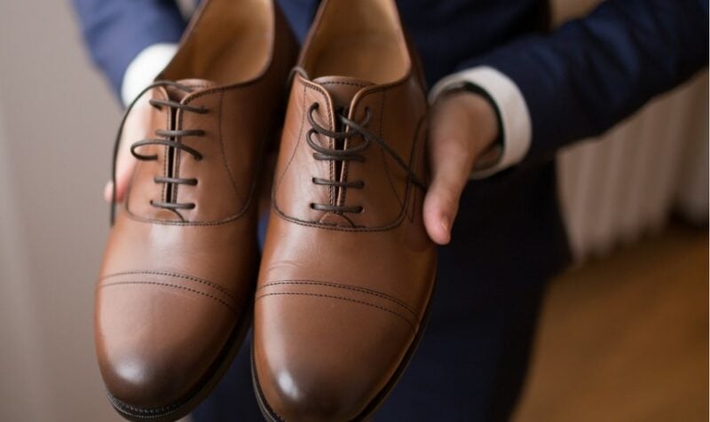 Man in a blue suit holding brown shoes and pointing them to the camera